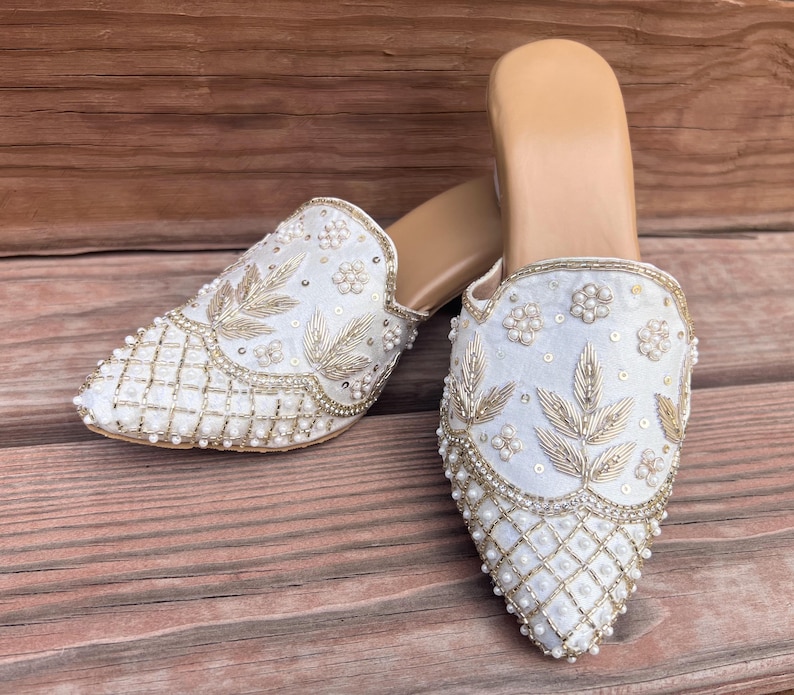 May include: A pair of white, slip-on mules with intricate embroidery and beadwork. The shoes feature gold-colored leaf and floral designs, with a grid pattern on the toe. The soles are a light tan color, and the shoes are set against a wooden background.