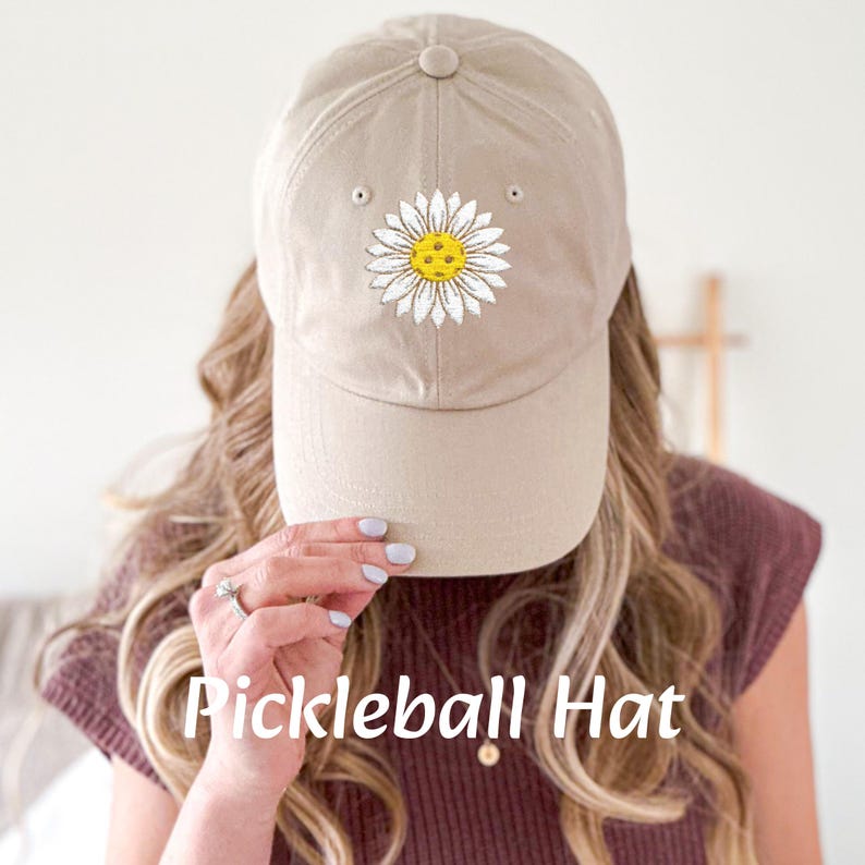 Puede incluir: Una gorra de b&eacute;isbol beige con una margarita blanca bordada en la parte delantera. La margarita tiene un centro amarillo con un dise&ntilde;o de pickleball.
