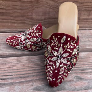 May include: Burgundy velvet slip-on shoes with intricate floral embroidery in gold and silver thread. The design features a central flower and leaf patterns, with small beads adding detail. The shoes have a neutral-colored insole.