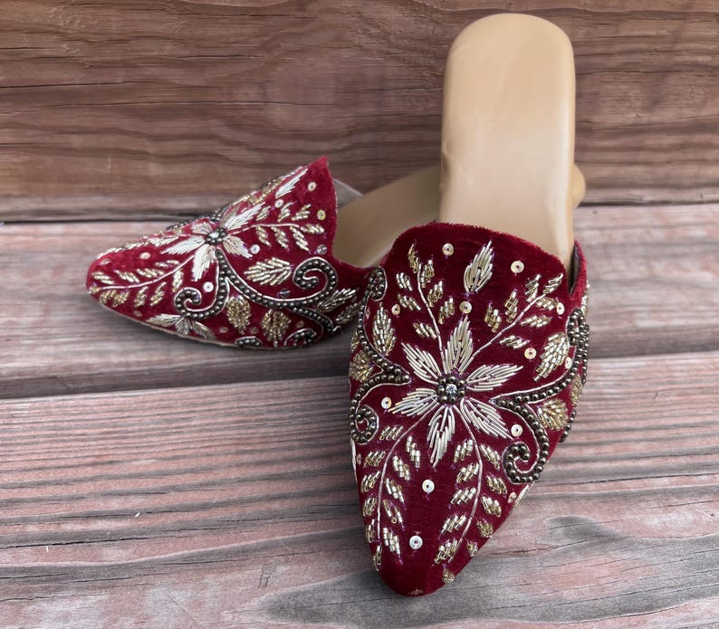 May include: Burgundy velvet slip-on shoes with intricate floral embroidery in gold and silver thread. The design features a central flower and leaf patterns, with small beads adding detail. The shoes have a neutral-colored insole.