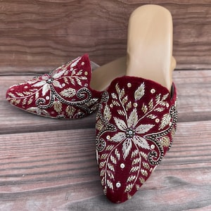 May include: A pair of burgundy velvet mules with intricate floral embroidery in gold and silver thread. The open-back shoes feature a pointed toe and are displayed on a wooden surface.