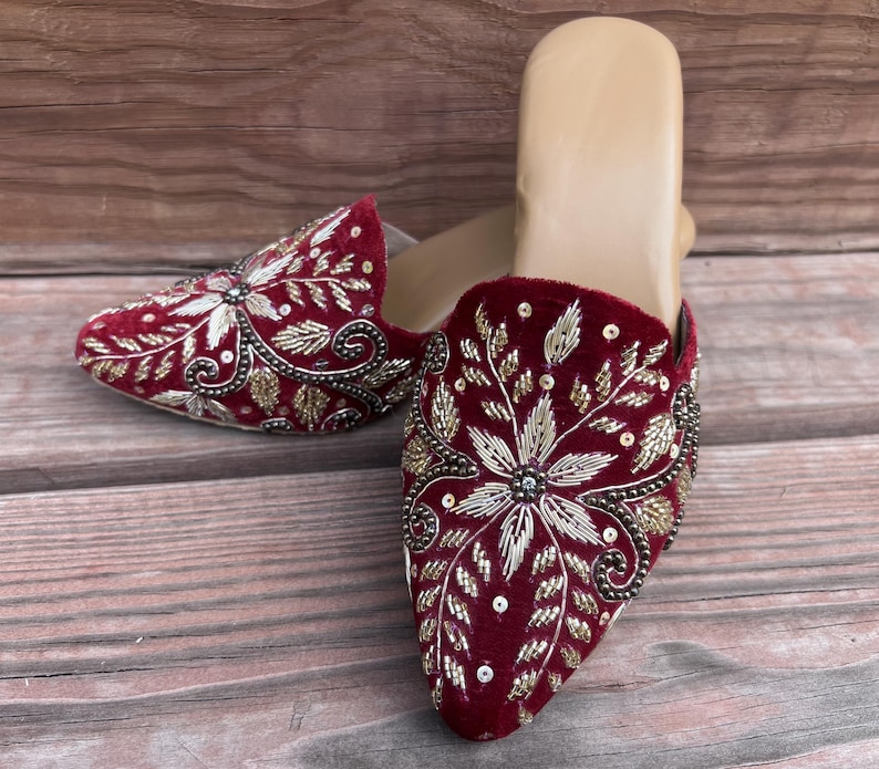 May include: A pair of burgundy velvet mules with intricate floral embroidery in gold and silver thread. The open-back shoes feature a pointed toe and are displayed on a wooden surface.