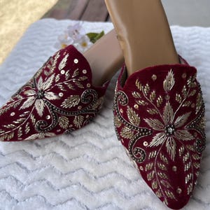 May include: A pair of burgundy velvet mules with intricate gold embroidery. The pointed-toe shoes feature floral and leaf patterns, embellished with sequins and beads. The shoes are on a white surface.