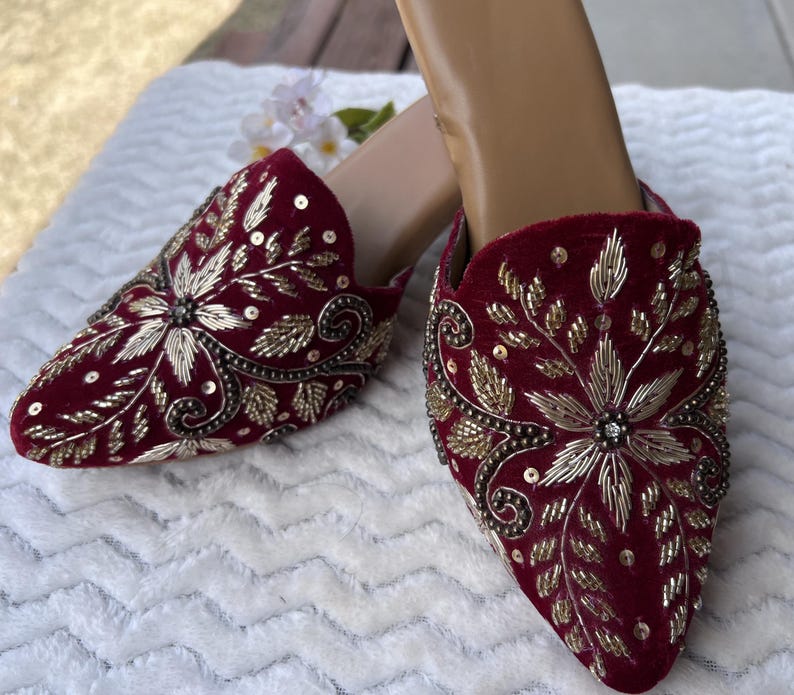 May include: A pair of burgundy velvet mules with intricate gold embroidery. The pointed-toe shoes feature floral and leaf patterns, embellished with sequins and beads. The shoes are on a white surface.
