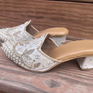 May include: A pair of ivory-colored, embellished mules with a low heel. The shoes feature intricate beadwork and embroidery with gold and pearl accents. The soles are a light tan color.