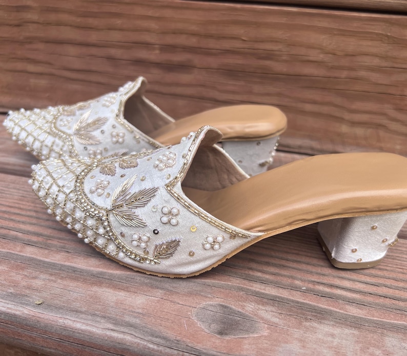 May include: A pair of ivory-colored, embellished mules with a low heel. The shoes feature intricate beadwork and embroidery with gold and pearl accents. The soles are a light tan color.