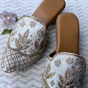 May include: A pair of ivory-colored slip-on shoes with intricate embroidery. The shoes feature gold-colored leaf and floral designs, along with pearl accents. The soles are a light brown color.
