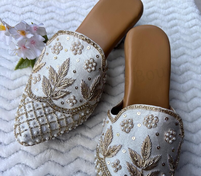 May include: A pair of ivory-colored slip-on shoes with intricate embroidery. The shoes feature gold-colored leaf and floral designs, along with pearl accents. The soles are a light brown color.