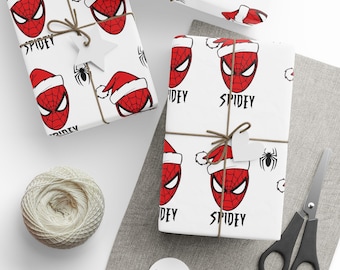 May include: White wrapping paper featuring a red Spider-Man face wearing a Santa hat, with the word "SPIDEY" in black. The gifts are tied with twine and adorned with star-shaped tags. A spool of twine, scissors, and tags are also present.