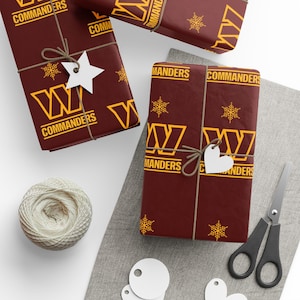 May include: Gift-wrapped presents with burgundy paper featuring the word "COMMANDERS" in gold. The gifts are tied with twine and adorned with white star and heart-shaped tags. Gold snowflakes and the team's logo are printed on the wrapping paper.