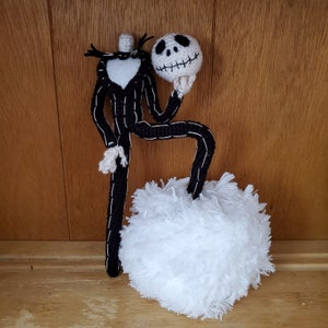 Op de afbeelding: Een zwart-witte gehaakte Jack Skellington pop die een wit gehaakt Jack Skellington hoofd vasthoudt. De pop staat op een witte, pluche wolk.