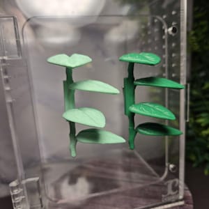May include: Two green plant stem sculptures with leaf-shaped shelves are displayed inside a clear acrylic box. The leaves are various shades of green. The box is partially open, revealing a glimpse of a green plant in the background.