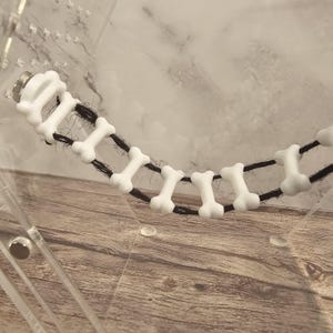 May include: A white plastic bone-shaped toy for pets, strung together with black string. The toy is in a clear plastic container.