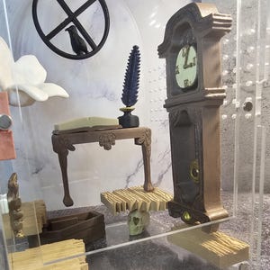 May include: A miniature room display with a wooden table, a clock, a skull, and a book. The room is enclosed in a clear acrylic case.