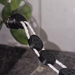 Peut inclure: Une cha&icirc;ne de boutons noirs &agrave; cinq trous chacun, reli&eacute;s par une ficelle blanche. Les boutons sont fix&eacute;s &agrave; une surface transparente.