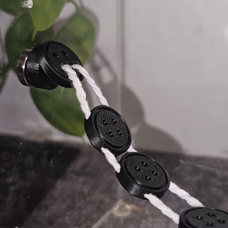 Peut inclure: Une cha&icirc;ne de boutons noirs &agrave; cinq trous chacun, reli&eacute;s par une ficelle blanche. Les boutons sont fix&eacute;s &agrave; une surface transparente.