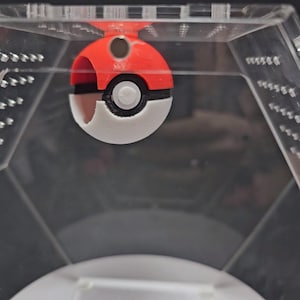 Puede incluir: Un juguete Pokeball rojo y blanco con un centro negro y un peque&ntilde;o bot&oacute;n blanco. El Pokeball est&aacute; dentro de un contenedor de pl&aacute;stico transparente.