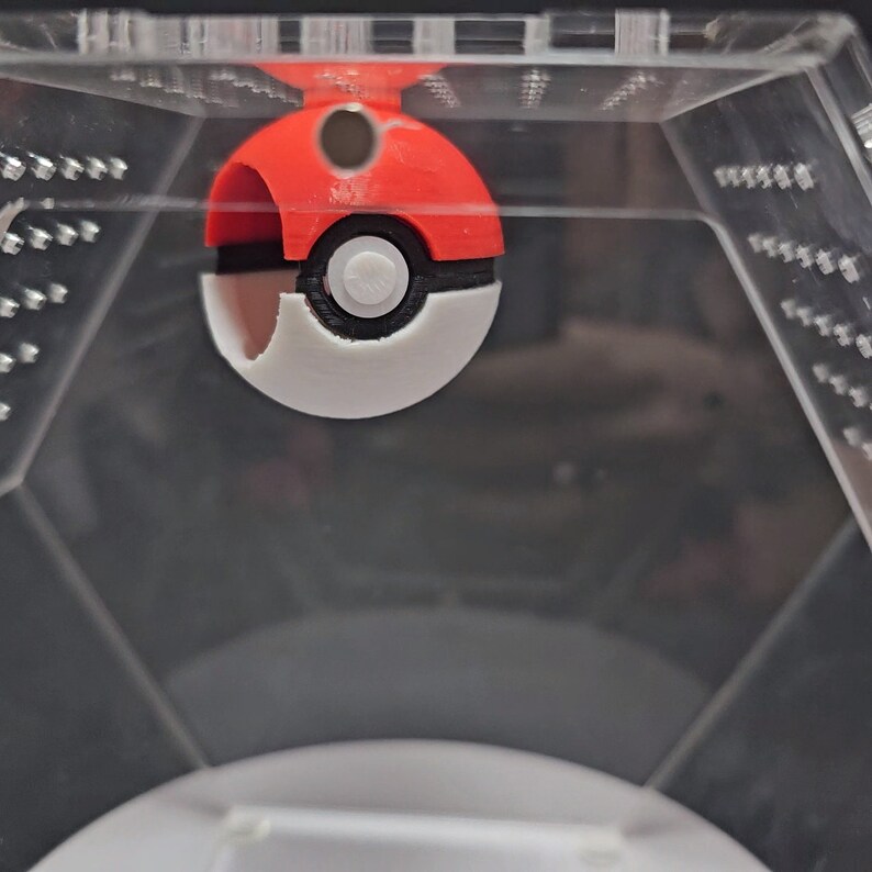 Puede incluir: Un juguete Pokeball rojo y blanco con un centro negro y un peque&ntilde;o bot&oacute;n blanco. El Pokeball est&aacute; dentro de un contenedor de pl&aacute;stico transparente.