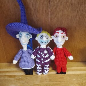 Pode incluir: Três bonecas de crochê inspiradas nos personagens do filme "O Estranho Mundo de Jack". A boneca à esquerda está usando um vestido roxo e um chapéu roxo alto. A boneca do meio está usando uma fantasia de esqueleto roxo e branco. A boneca à direita está usando um vestido vermelho.