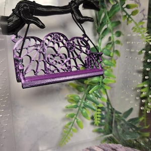 May include: A miniature purple swing with an intricate web-like backrest featuring a central heart shape. The swing is suspended by black cords from a black, textured branch-like structure, creating a hanging display piece for small figures.