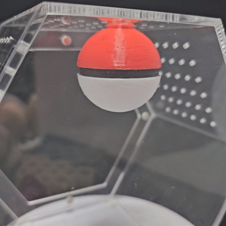 Puede incluir: Una esfera roja y blanca con una banda negra alrededor del medio cuelga de la parte superior de un contenedor hexagonal de pl&aacute;stico transparente.