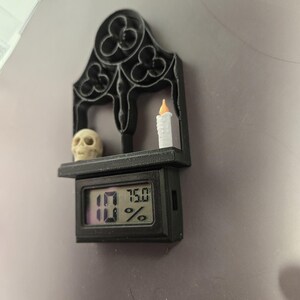 以下が含まれることがあります： 黒いゴシック様式の棚。頭蓋骨、ろうそく、10％と75.0％を表示するデジタルディスプレイがあります。棚は装飾的な切り欠きのあるアーチ型のトップを持っています。ろうそくはオレンジ色の炎です。