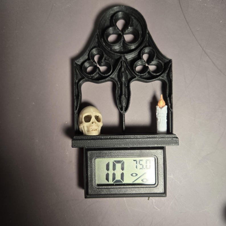 以下が含まれることがあります： 黒いゴシック様式の棚。骸骨、ろうそく、デジタル表示があります。表示は10と75.0％を示しています。棚はアーチ型のトップと装飾的な切り抜きがあります。骸骨はオフホワイトで、ろうそくはオレンジ色の炎が付いた白です。