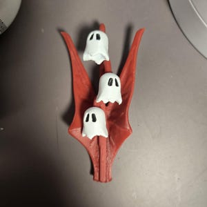 May include: A 3D-printed Halloween decoration. It features three white ghost figures with black eyes, mounted on a red, stylized wing-shaped base. The ghosts have a simple, cartoonish design, and the base has a textured surface.