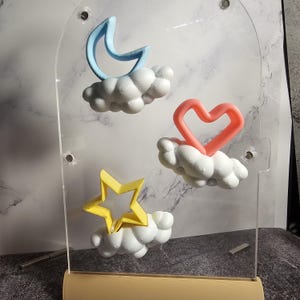 May include: A clear acrylic display stand with three 3D printed shapes: a blue crescent moon, a red heart, and a yellow star. Each shape is attached to a white cloud-shaped base.