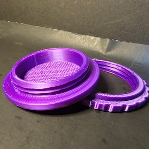 May include: A purple 3D printed container with a lid. The container has a grid pattern on the bottom and a textured rim. The lid has a textured rim and a smooth top.