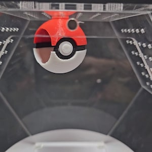 Puede incluir: Un juguete Pokeball rojo y blanco con un centro negro y un bot&oacute;n blanco. El Pokeball est&aacute; colgado del techo de una caja de pl&aacute;stico transparente.