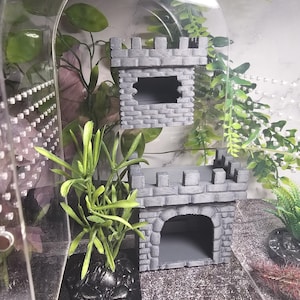 May include: Two grey castle-shaped reptile hides with arched openings. The hides are made of a textured material that resembles stone. The hides are placed on a grey and white textured surface.