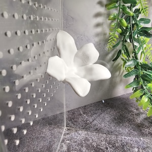 May include: A white ceramic flower with five petals is attached to a clear plastic surface. The flower is in the corner of the image, and there is a green plant in the background.