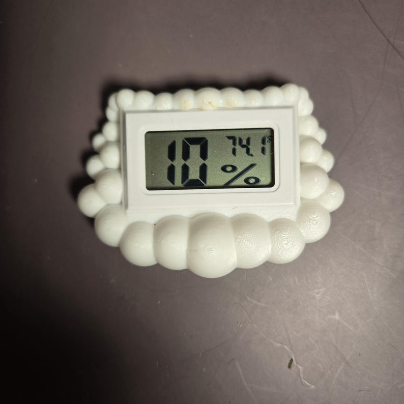 Peut inclure: Un thermom&egrave;tre num&eacute;rique blanc avec une bordure en forme de bulles affiche les chiffres "10" et "74,1%" sur un &eacute;cran noir. L'appareil est con&ccedil;u pour mesurer la temp&eacute;rature et l'humidit&eacute;.