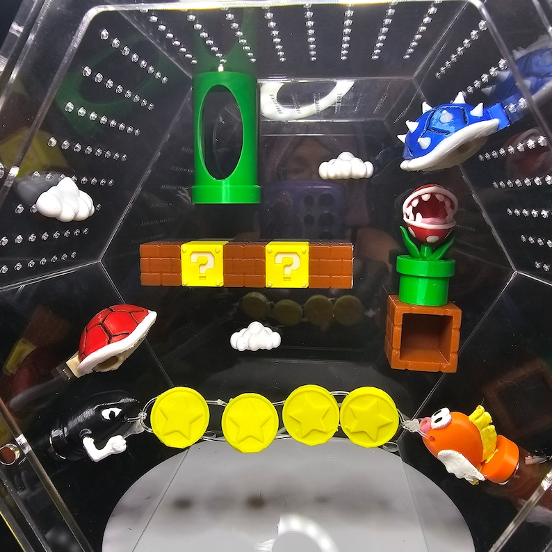 May include: A collection of Super Mario Bros. themed figurines, including a green pipe, a question block, a koopa troopa, a piranha plant, a goomba, a cheep cheep, and gold coins. The figurines are displayed in a clear plastic hexagonal container.