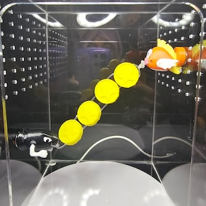 May include: A yellow and orange toy fish with a white string attached to it. The string is connected to a series of six yellow plastic discs. A black toy figure with a white string attached to it is on the left side of the image.