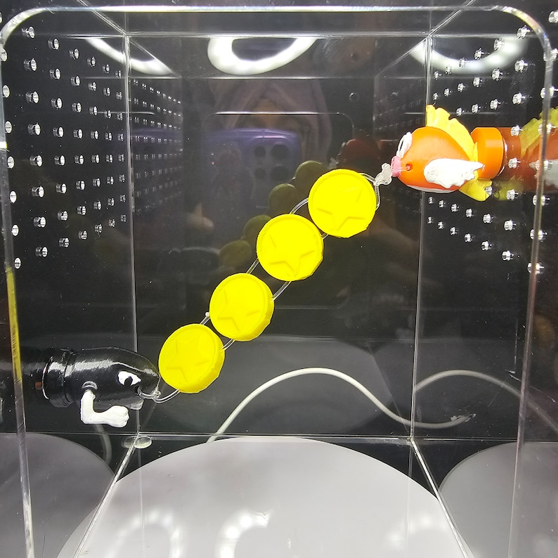 May include: A yellow and orange toy fish with a white string attached to it. The string is connected to a series of six yellow plastic discs. A black toy figure with a white string attached to it is on the left side of the image.