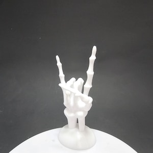 May include: White skeletal hand figurine making the rock and roll hand gesture. The hand is on a white base.
