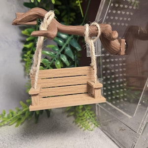 May include: A miniature wooden swing in light brown, suspended by twine ropes from a brown branch. The swing has a slatted seat. It is set against a backdrop of green foliage and a clear plastic enclosure.