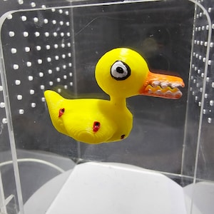 May include: A yellow rubber duck with red spots and a large orange beak with white teeth. The duck is in a clear plastic container.