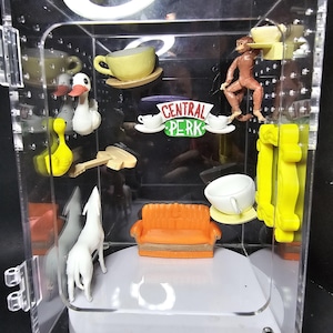 May include: A clear acrylic display case with various miniature figurines and props, including a couch, a coffee cup, a monkey, and a sign that reads "Central Perk".