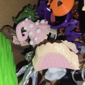 May include: A selection of 3D-printed Halloween decorations. Features an orange jack-o'-lantern, a pink mushroom with white spots, and other colourful, whimsical shapes. The items are in a cardboard box.
