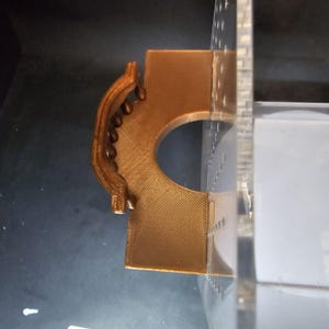 Puede incluir: Una pieza decorativa de pl&aacute;stico marr&oacute;n impresa en 3D con un dise&ntilde;o curvo y abierto. La pieza est&aacute; unida a un panel de acr&iacute;lico transparente.
