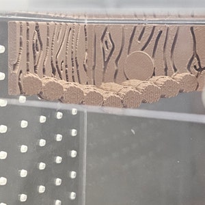 May include: A close-up of a 3D printed object with a wood grain texture. The object is brown and has a circular shape in the center. The object is sitting on a clear surface.