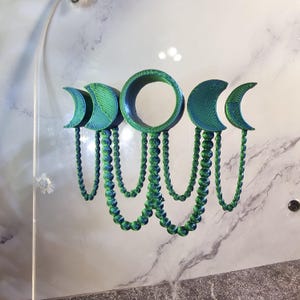 May include: A green and blue 3D printed moon phase wall hanging with a beaded chain. The moon phases are in a crescent shape and the beads are round.