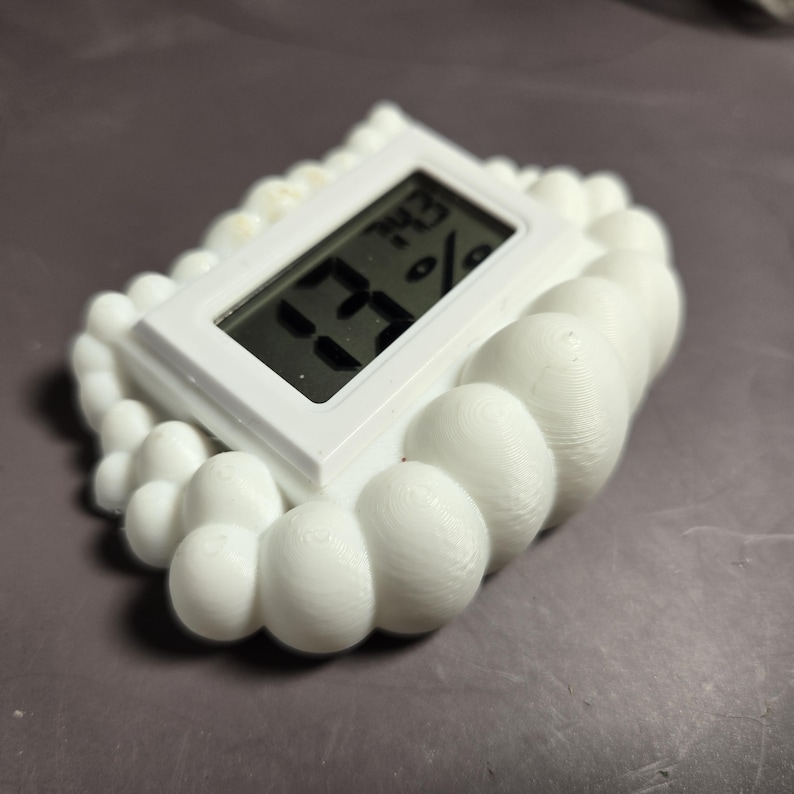 Peut inclure: Un hygrom&egrave;tre num&eacute;rique blanc avec un cadre en forme de nuage affiche 73% et 74,3. L'appareil est rectangulaire avec des bords arrondis et un petit &eacute;cran LCD. Le cadre a un design textur&eacute; et bulleux.