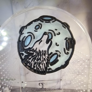 May include: A black and white 3D printed design of a wolf howling at the moon. The moon is light blue and has a textured surface.