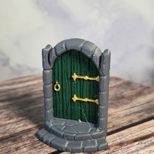 Op de afbeelding: Een miniatuur fantasiedeur met een groene deur en gouden hardware. De deur is geplaatst in een grijze stenen lijst met een stenen voet. De deur is een grillig decoratief item.