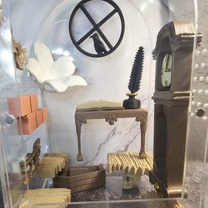 May include: A miniature dollhouse room with a wooden desk, a book, a quill pen, an inkwell, a clock, a coffin, and a skull. The room is decorated with a white flower, a brick wall, and a wooden floor.