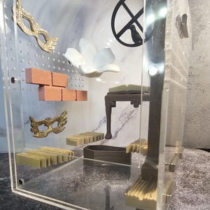 May include: A clear acrylic box with a variety of miniature objects inside, including a white flower, a brick wall, a small table, and two gold masks. The box is sitting on a marble surface.